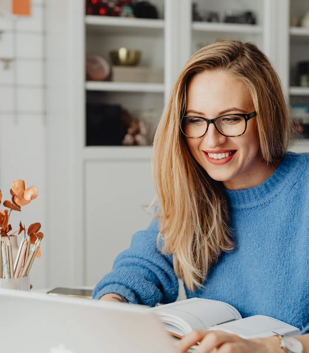 Femme aux cheveux blonds, souriante, assise devant son ordinateur à son bureau de travail, feuilles orangées dans un vase en verre